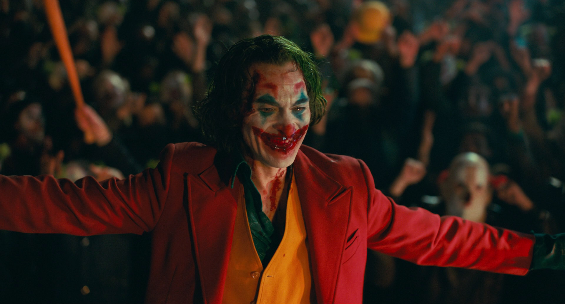 the image shows the joker, played by joaquin phoenix, in his iconic red suit and green hair. he is surrounded by a chaotic crowd of people, and his face is painted with a grotesque, sinister smile. the scene is dark and unsettling, reflecting the character's madness and chaos. the joker is a popular character from dc comics, and this image captures his essence perfectly.