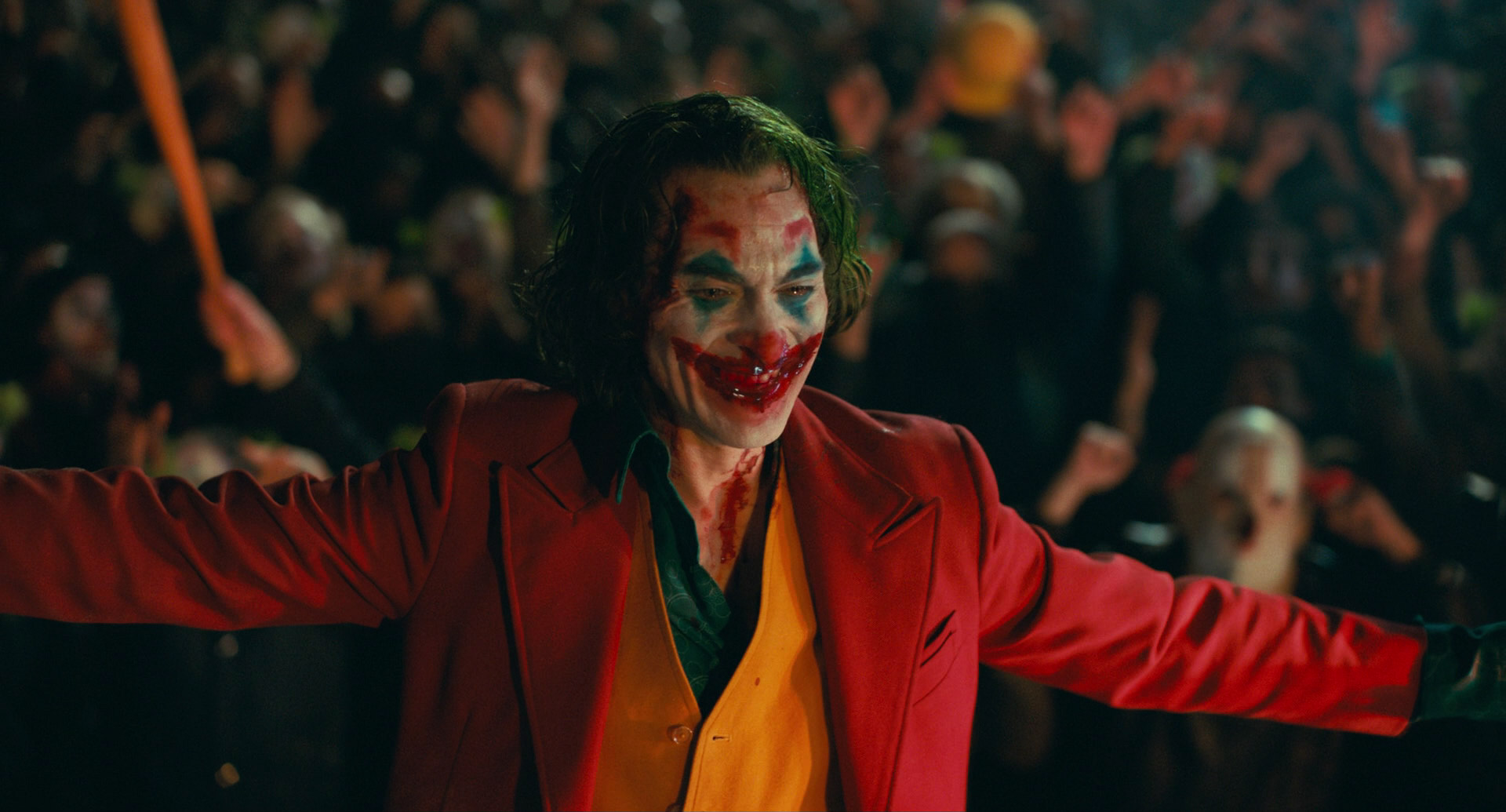 a still from the movie joker, showing joaquin phoenix as the joker.  he is in full costume and makeup, standing with his arms outstretched towards the viewer, a wide, manic grin on his face. he is surrounded by a large, cheering crowd in the background, with the scene lit by colorful artificial lights that create a sense of chaos and excitement.