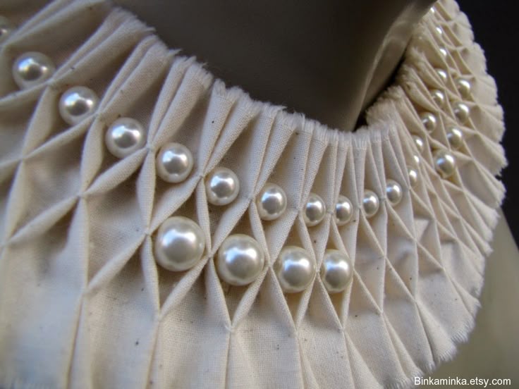 a closeup shot of a fabric collar adorned with pearls. the offwhite fabric is pleated in a geometric diamond pattern, with a pearl placed at each intersection. the collar is intricately crafted, suggesting a handmade, couture piece of fashion or textile art.