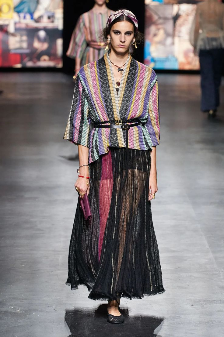 a fashion model walks the runway in a stylish outfit. she is wearing a colorful striped jacket over a black sheer skirt, a black belt, and black flats. the model also has a headband, jewelry, and a bag. she has long brown hair and is wearing makeup. the runway is a modern, minimalist space.
