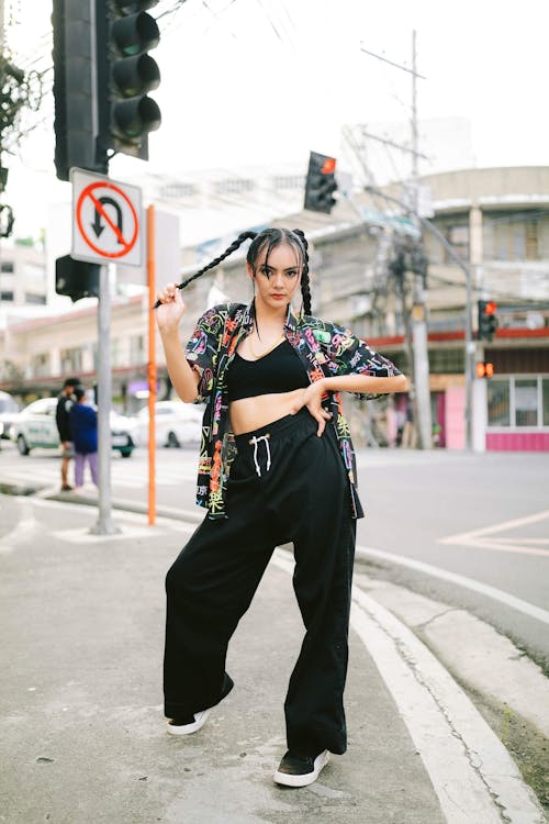 a young woman with pigtails is standing confidently on a city street.  she's wearing black wideleg pants, black sneakers, and a colorful graffitiprinted shirt over a black crop top. her pose is relaxed yet stylish, showcasing her urban streetwear fashion. the background features city buildings, traffic lights, and a crosswalk. the overall atmosphere is one of contemporary urban cool.