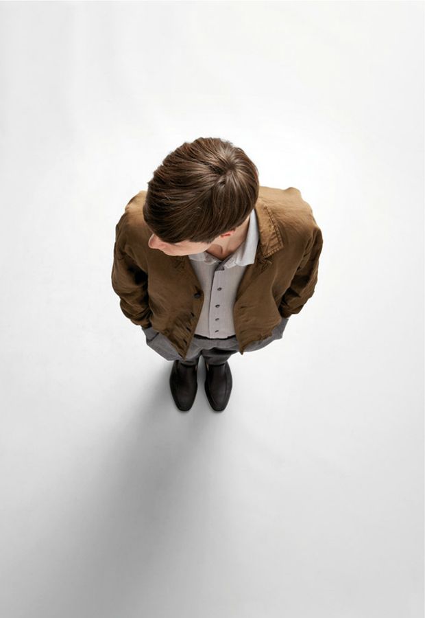 a man is standing on a white surface with his head looking down. he is wearing a brown jacket, a white shirt, gray pants, and black shoes.