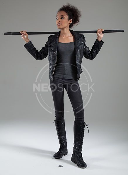 a young woman stands facing away from the camera in a studio setting.  she is wearing a black leather jacket with a black tshirt, black skinny jeans, and long black boots. she is holding a black pole or stick.