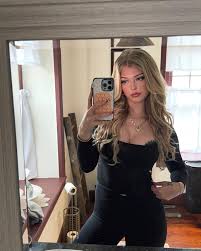 a woman is taking a mirror selfie in a bathroom. she has blonde hair and is wearing a black outfit with a necklace. she is holding a smartphone in her hand.