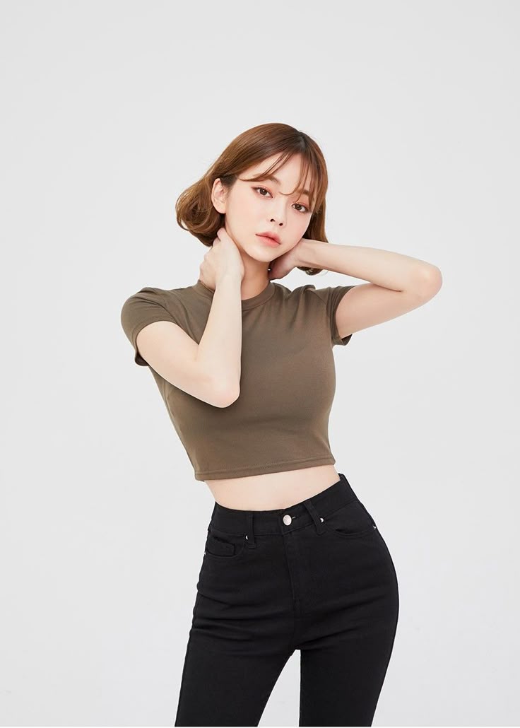 a young woman with short brown hair is posing in front of a white background. she is wearing an olive green crop top and highwaisted black jeans. her hands are behind her neck.