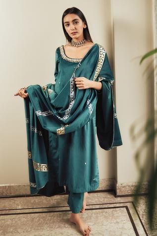 a fulllength shot of a woman modeling a teal green silk kurta and shalwar kameez with gold and silver embroidery. the outfit includes a matching dupatta draped over her shoulders.  the woman is posing indoors against a neutral background with a visible part of a wall and some plants in the background.  she is barefoot. the overall style is elegant and traditional south asian fashion.