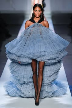 a woman walks the runway in a stunning light blue tulle gown with a high low skirt, accessorizing with black heels and silver earrings. her hair is pulled back and she is walking gracefully in a welllit runway with white tile.
