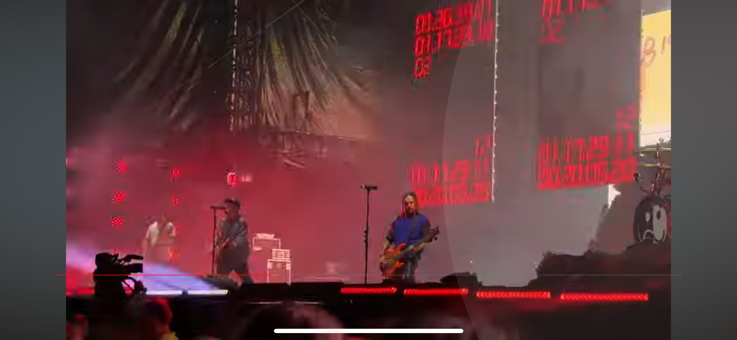 a blurry image of a band performing on stage in front of a crowd. the stage is lit with red lights and fog is present. the band is playing guitars and singing. the drummer is in the back of the stage behind a drum set. the crowd is silhouetted in the background. the image is taken from a low angle looking up at the stage.