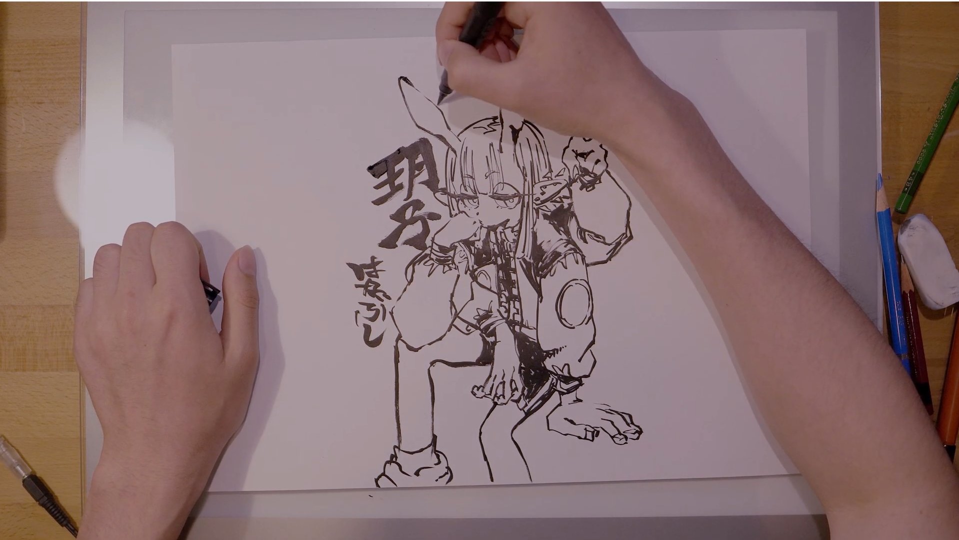 the image shows an artist's hands drawing a mangastyle character with a black marker on a bright drawing board.  the drawing board is placed on a table and we see drawing tools, pencils and markers next to it.  the image is taken from above.