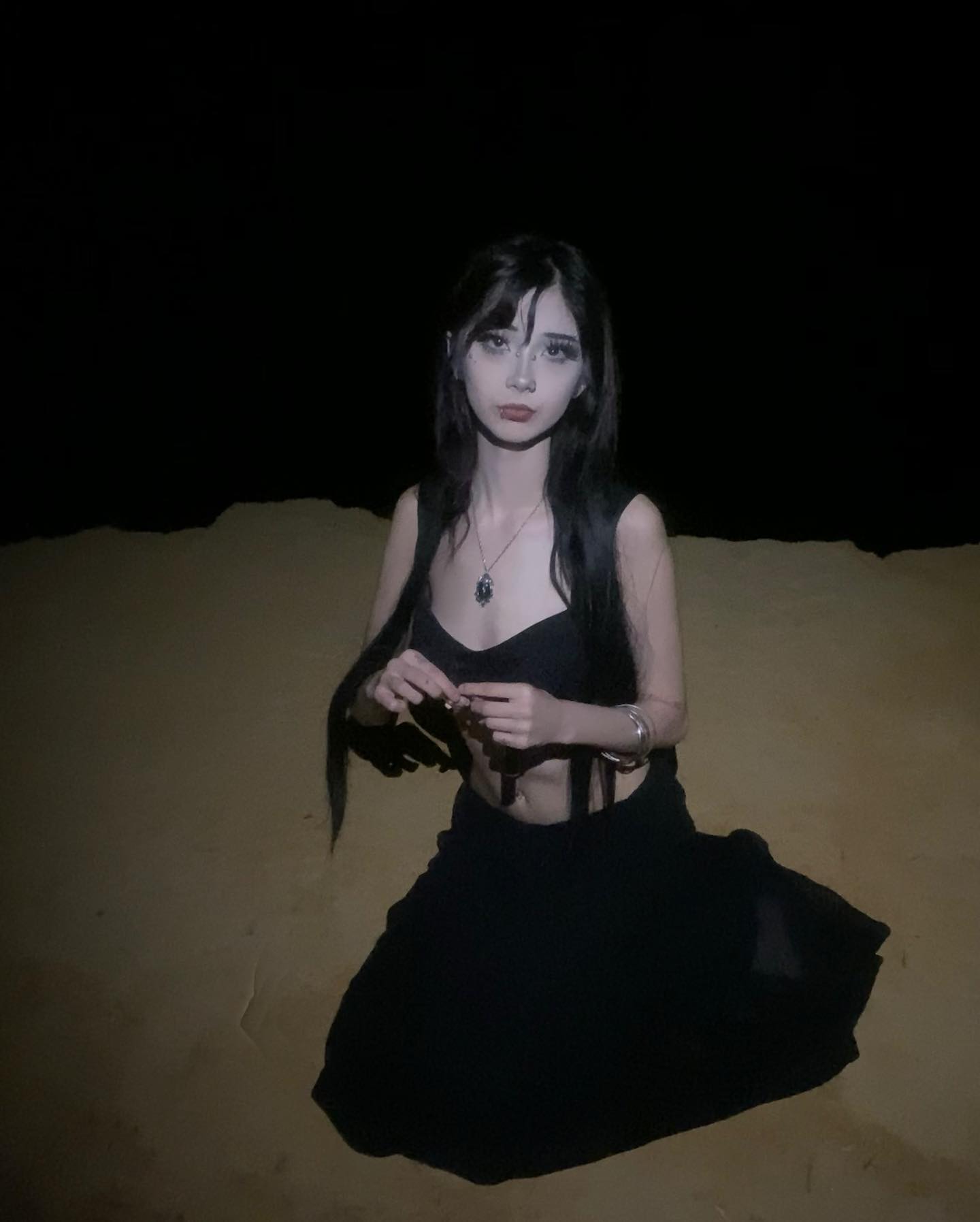 a young woman with long dark hair and dark attire is posed in the desert at night.  she is illuminated by artificial light, creating a dramatic lowkey lighting effect. the overall aesthetic leans towards a goth or alternative style.