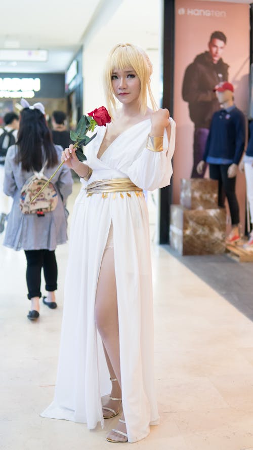 a young woman dressed in a white cosplay dress, holding a red rose. she has blonde hair and is standing in a shopping mall.  the background features mannequins and displays. the overall impression is of a character cosplay, possibly from an anime or manga.