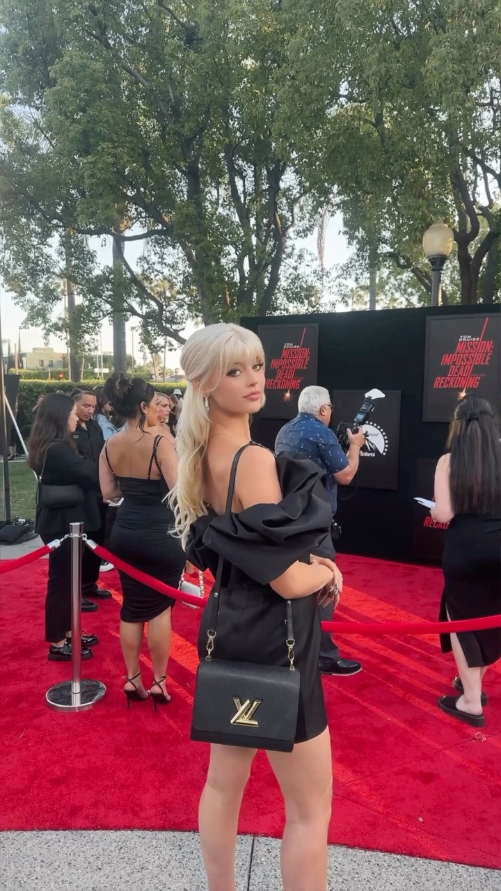 a woman in a black dress poses for a photo on a red carpet. she is carrying a louis vuitton bag. behind her are several people, including a photographer and a building with trees in front of it. the red carpet is for the premiere of mission impossible dead reckoning part one.