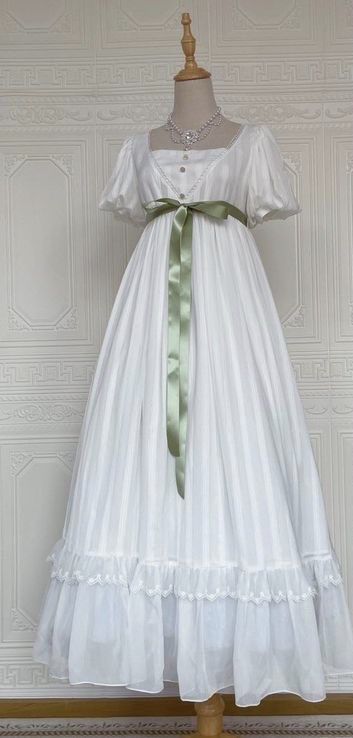 a long white dress with a green ribbon is on display against a white wall with subtle patterns. the dress has short puff sleeves, a square neck, an empire waist, and a flowing skirt with lace trim. the soft lighting creates a romantic and feminine atmosphere.