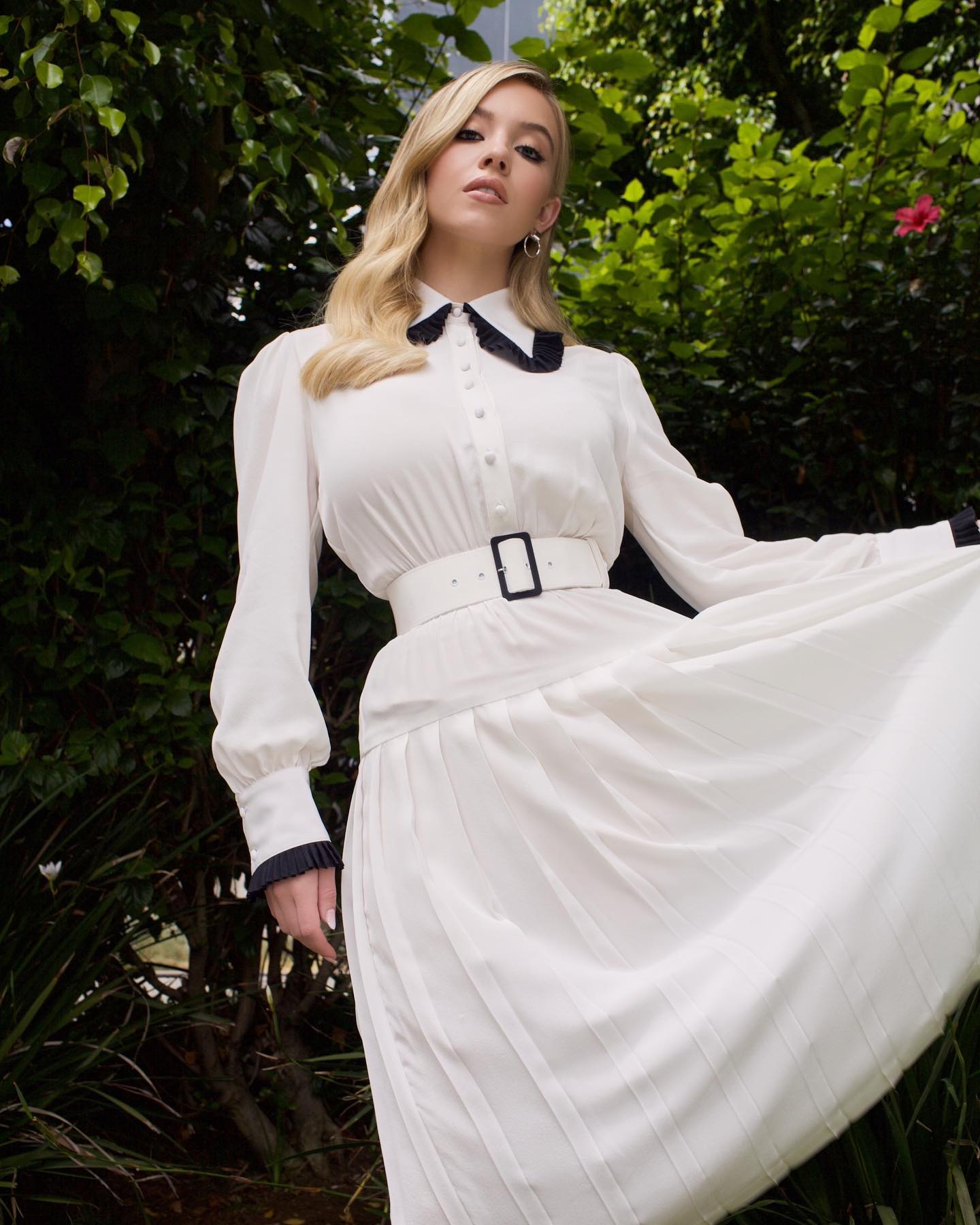 the image shows a young blonde woman wearing a stylish, longsleeved white dress. the dress features a pleated skirt, a black peter pan collar, and black cuffs. a simple white belt cinches the waist. the model is posed outdoors in a lush green garden setting. her expression is confident and slightly serious. the overall aesthetic is elegant and hints at a vintage or retro style.