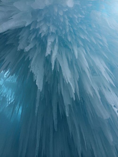 the image shows a closeup view of numerous icicles hanging from a ceiling. the icicles are various sizes, with some short and stubby and others long and slender. the ice has a blue hue, creating a cold and wintery atmosphere. the perspective is looking upwards, emphasizing the dense formation of icicles.
