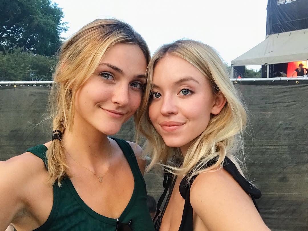 a closeup selfie of two young blonde women, smiling happily at the camera. they appear to be at an outdoor summer music festival, with a dark tarp and stage visible in the background. one woman wears a green tank top, the other a black top.  they are both casually dressed and have their blonde hair loose.  the overall impression is one of carefree friendship and summer fun.