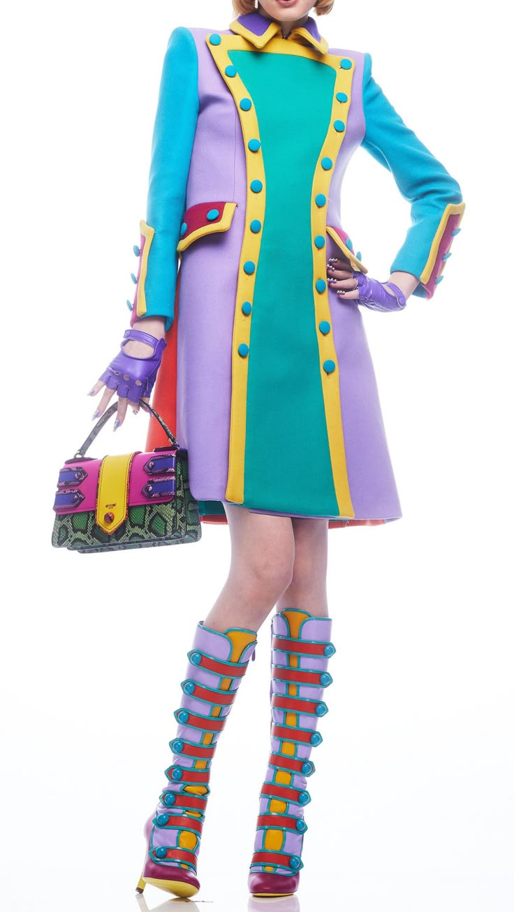 a woman is standing in a colorful outfit consisting of a coat, boots, purse, and fingerless gloves. the coat is purple with turquoise panels down the front and on the sleeves, outlined in yellow. it has buttons and flap details. the purse is rectangular and made of snake print material with pink and yellow panels. the boots are purple with yellow and red accents and button details. the woman is wearing fingerless gloves in purple. the background is white.