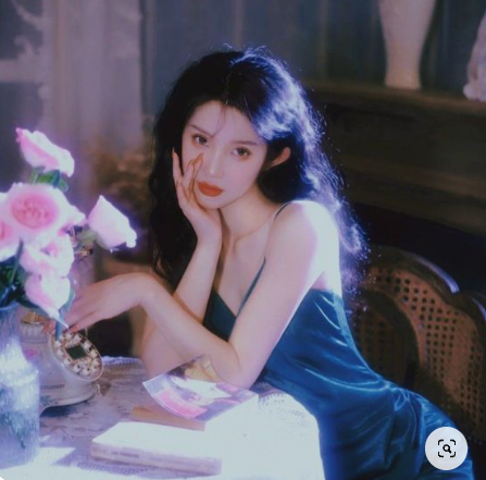 a young woman in a teal satin dress is seated at a table. she is leaning on her left hand and looking directly at the viewer. she has long black hair, red lips, and a vintage aesthetic. there is a bouquet of pink roses in a vase on the table, a rotary phone, and a magazine. the image has a soft, vintage filter applied.