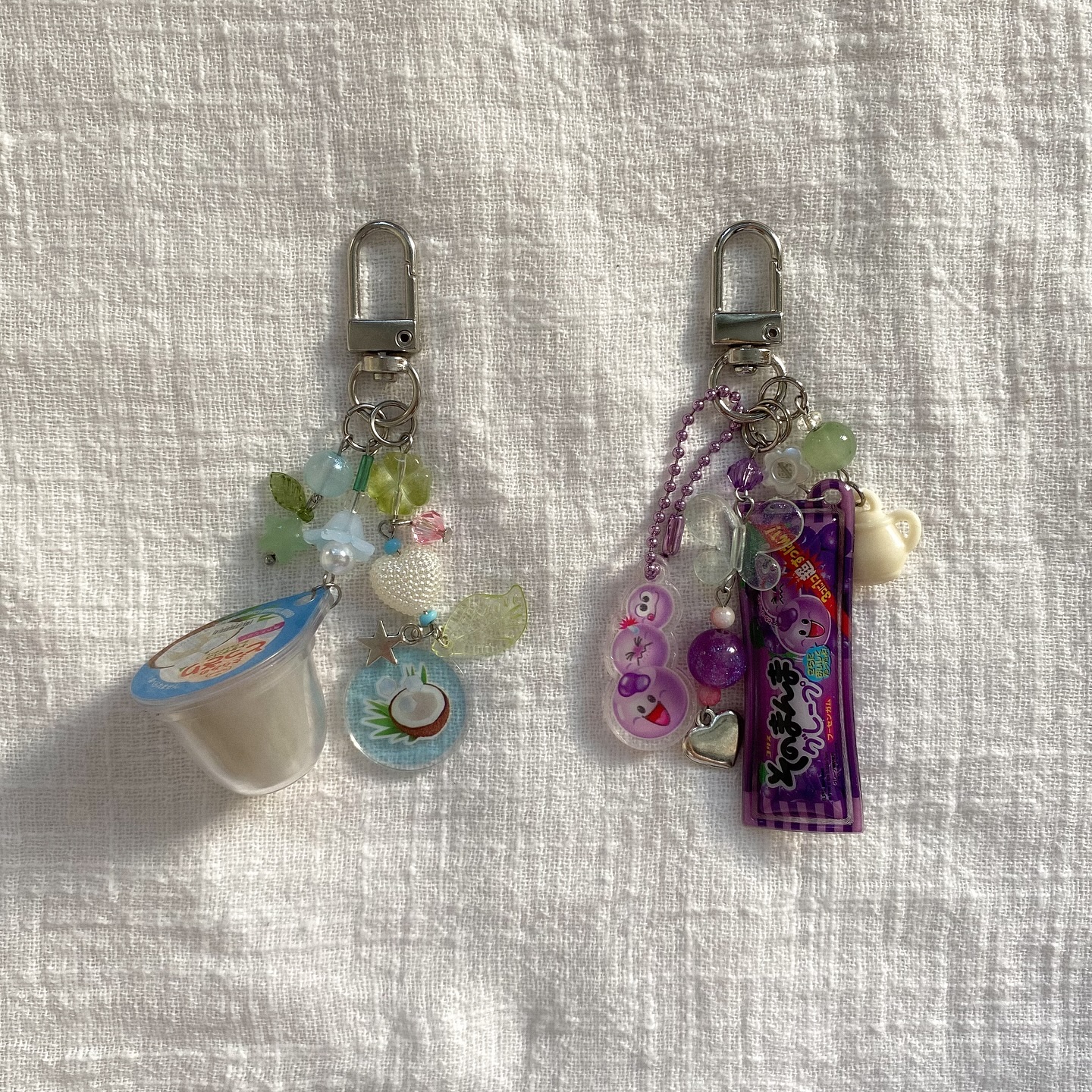 the image shows two handmade keychains lying on a white textured fabric. the keychain on the left has a small container resembling coconut pudding or yogurt, a clear charm with a coconut design, various beads in green, blue, and white, and silver clasp. the keychain on the right features a purple candy package charm, a clear charm with a cartoon character, purple beads, a silver heart charm, a tiny teapot charm and a silver clasp. both keychains have multiple small charms and beads attached, creating a whimsical and decorative effect.
