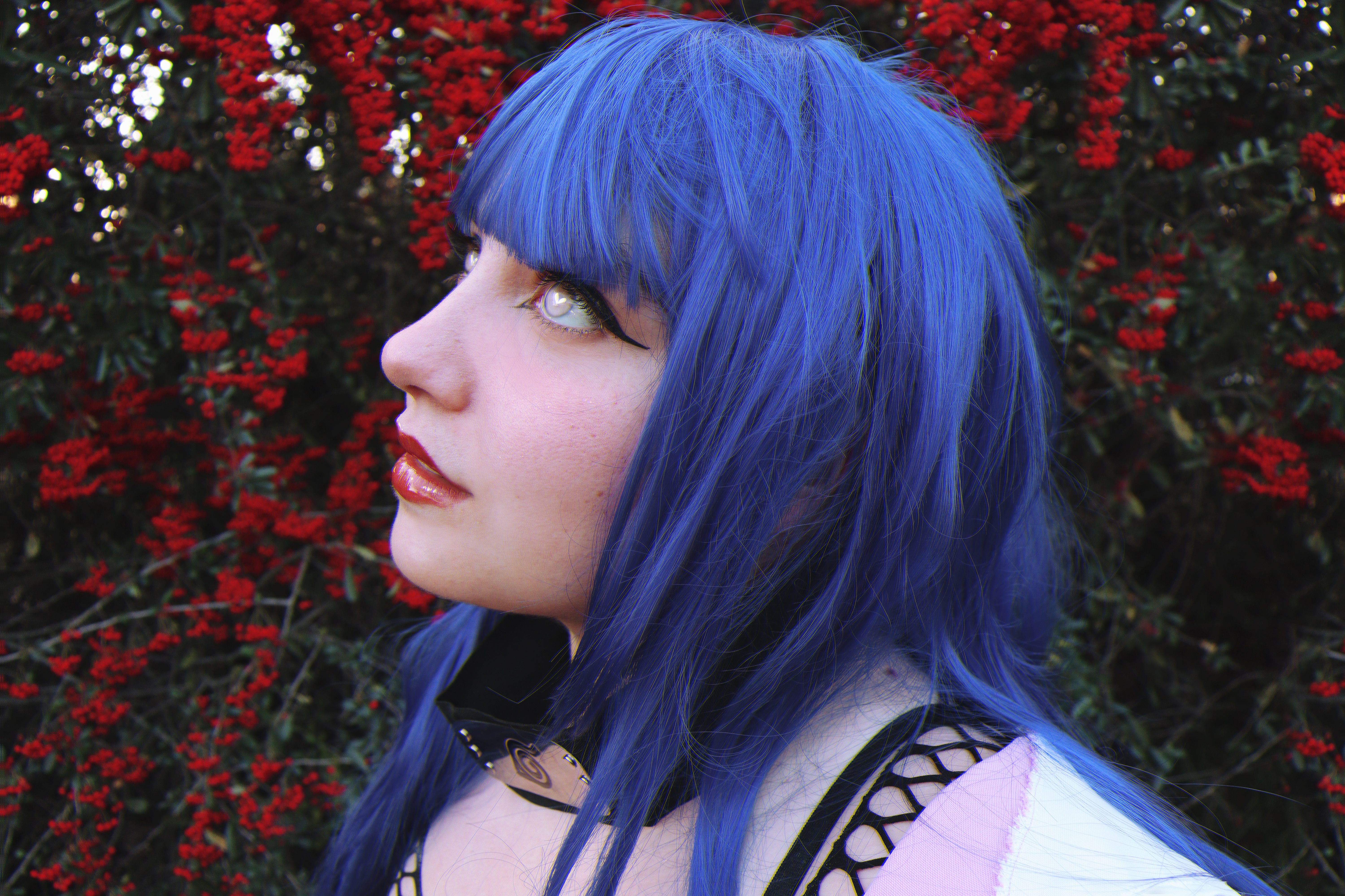 a person cosplaying as hinata hyuga from the anime naruto. she has a blue wig, white eyes, and is wearing a headband. she is standing in front of a bush with red flowers. the image is a side profile of her.