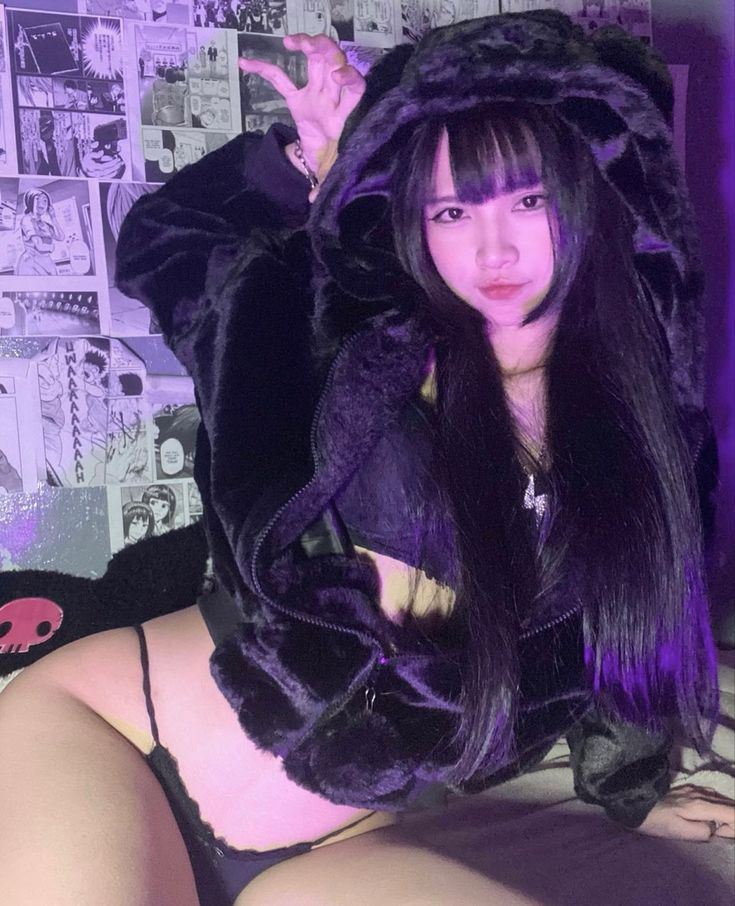 an asian woman is wearing black lingerie and a black furry hoodie with ears. she has long black hair with bangs. there are manga pages in the background.