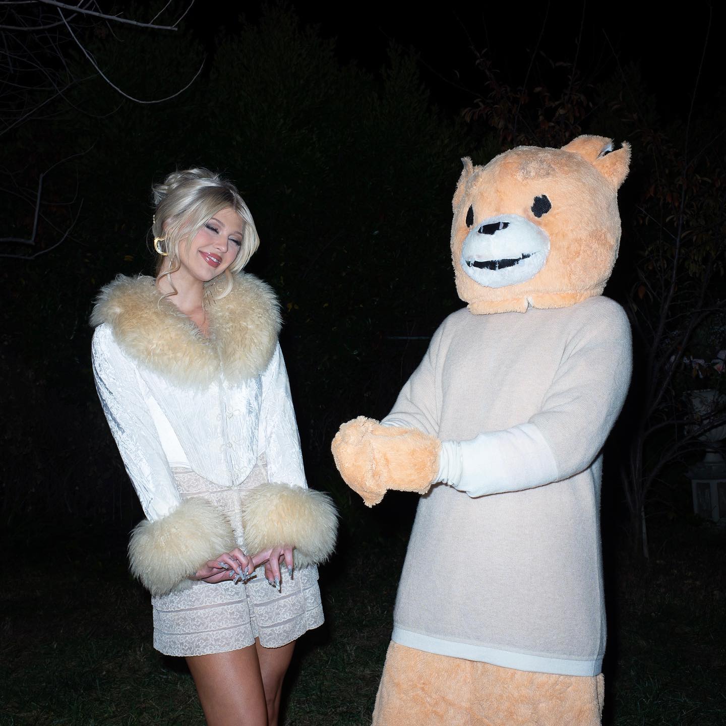 a woman with blonde hair is posing next to a person in a teddy bear costume. the woman is wearing a white jacket with a fur collar and fur cuffs, and short shorts. she has gold hoop earrings. the teddy bear costume is light brown and has a slightly creepy smile. they are standing outside with trees in the background and the scene is dark.