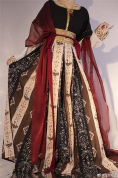 a woman is modeling a long, flowing dress. the dress is a mix of black, red, and cream fabrics with an intricate paisleylike pattern. it has long, wide sleeves and a cinched waist with a gold belt. the woman is shown from the waist down and is holding up the skirt of the dress slightly to show off the pattern. the background is a neutral beige color and the lighting is soft, giving the image a vintage feel.