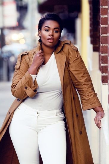 a woman is standing outdoors, wearing a white shirt, white jeans, and a long brown trench coat. she is leaning against a brick wall.