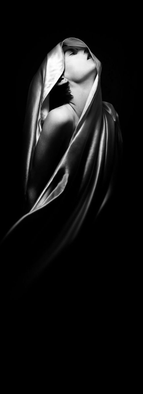 a black and white portrait of a woman with her head wrapped in a satin fabric. the fabric is draped over her head and shoulders, obscuring her features. her eyes are closed and she is looking up. the image is shot in a studio setting and the lighting is dramatic, creating deep shadows and highlights. the overall mood is sensual and mysterious.