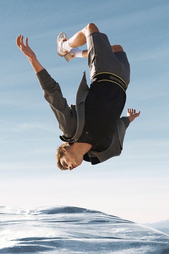 a young man wearing a black tshirt, a grey sweater, grey shorts, and calvin klein underwear, flips backwards in the air against a blue sky and white clouds. he is wearing white socks and white sneakers.