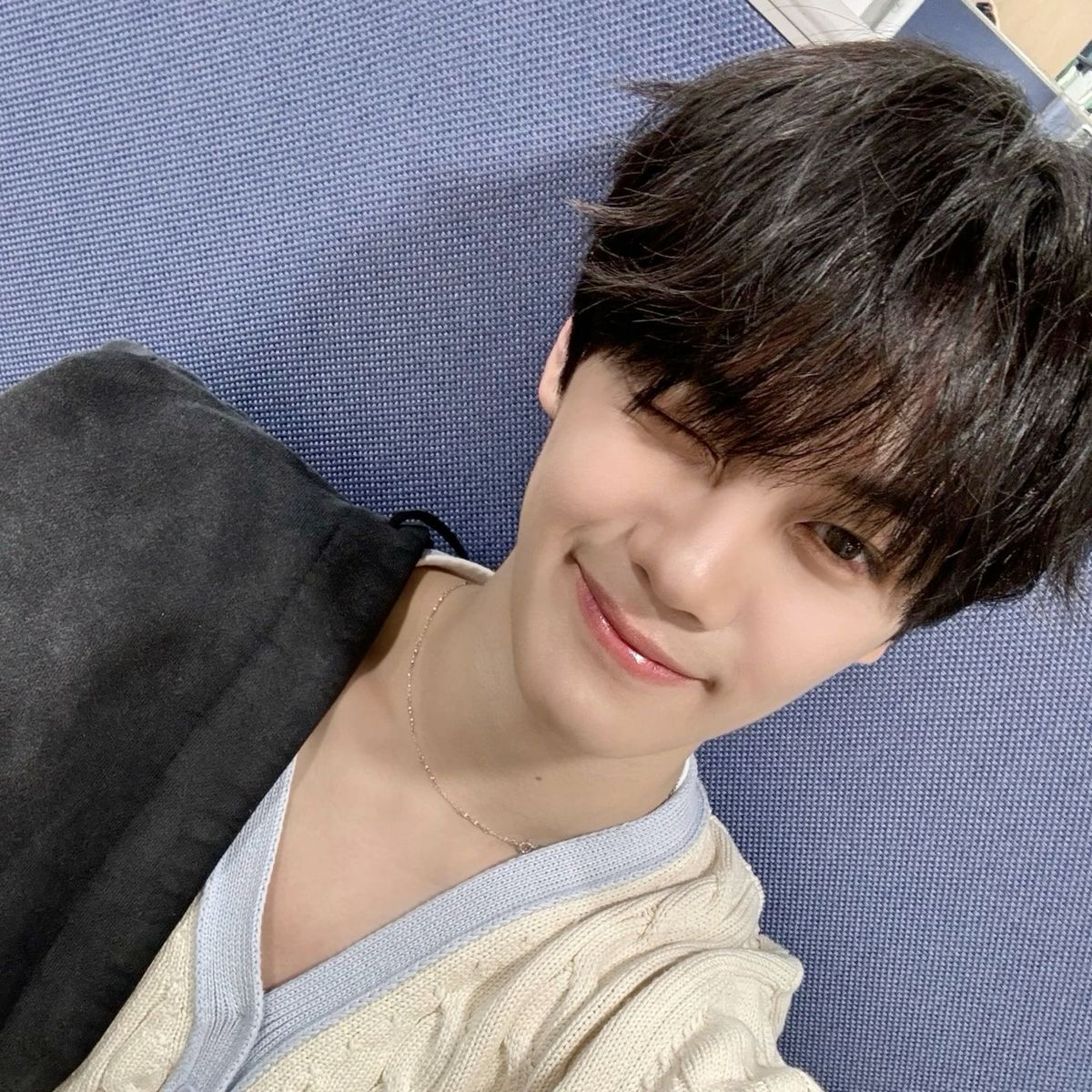 a closeup selfie of a young asian man with dark hair styled in a middle part. he is wearing a creamcolored knitted sweater and a thin silver chain. he has a soft smile on his face and is winking at the camera. the background is a blue textured surface. the lighting is soft and flattering, highlighting his features.