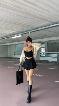 a woman is walking through a parking lot wearing a black skirt, white cardigan, and black boots. she's carrying a black bag and her hand is covering her face.