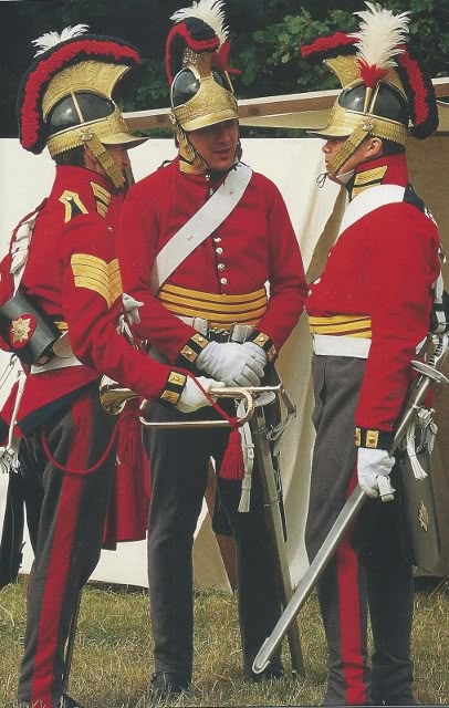 three soldiers in red british army uniforms stand with instruments and swords. they wear distinctive golden helmets with red and white plumes. one soldier holds a trumpet and another a sword. they are outdoors.