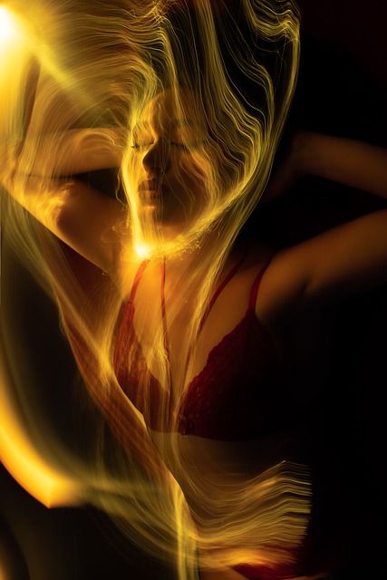 a woman's portrait with a flowing golden light painting around her. the light highlights her features, making her appear ethereal and mysterious. she is wearing a red bra and a seductive expression. the overall effect is dreamy, sensual and artistic.