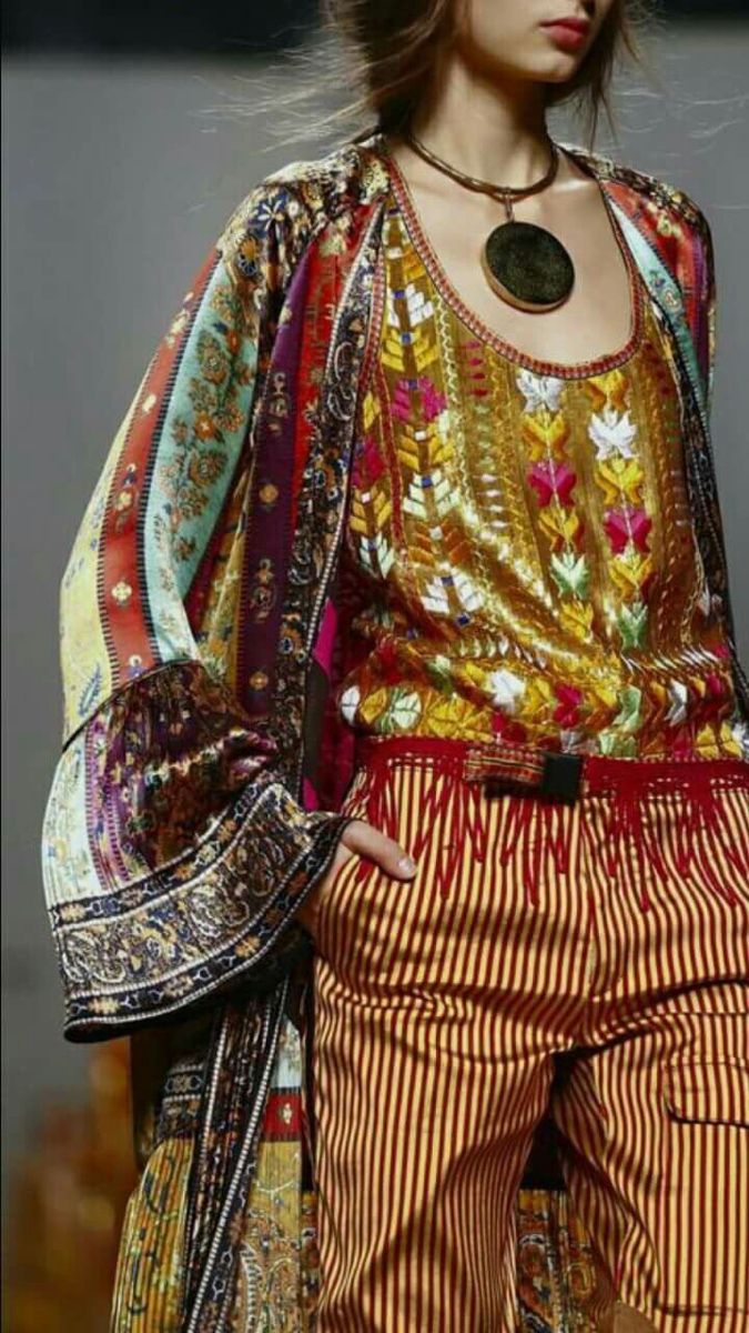 a woman is showcasing an eclectic fashion ensemble. she is dressed in an array of clothing items that include outerwear, a top, and pants. the clothing is adorned with vibrant patterns, textiles, and colors, giving it a bohemian and ethnic flair. a necklace and other jewelry enhance her style. the pants are striped, and the top appears to feature embroidery, while a belt with fringe accentuates her waist.