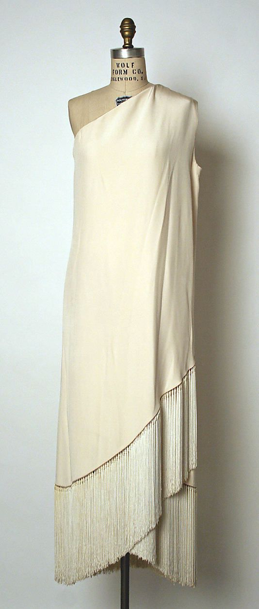 a white oneshoulder dress with fringe detailing on the skirt. the dress is made of silk and is a vintage style. the dress is displayed on a mannequin.