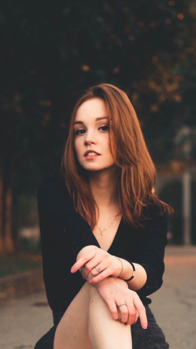 a young woman with long reddishbrown hair sits confidently in a park setting. she is wearing a black vneck top and has several pieces of jewelry on, including rings and a necklace.  the image has a soft, natural light, giving it a warm and inviting feel.