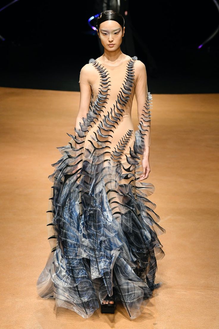 a woman in a  futuristic avant garde dress walks down a runway. the dress is  transparent  with intricate black lines and  layered  blue and black fabric.  the model is  wearing  a  hairpiece  and her  makeup  is  minimal.