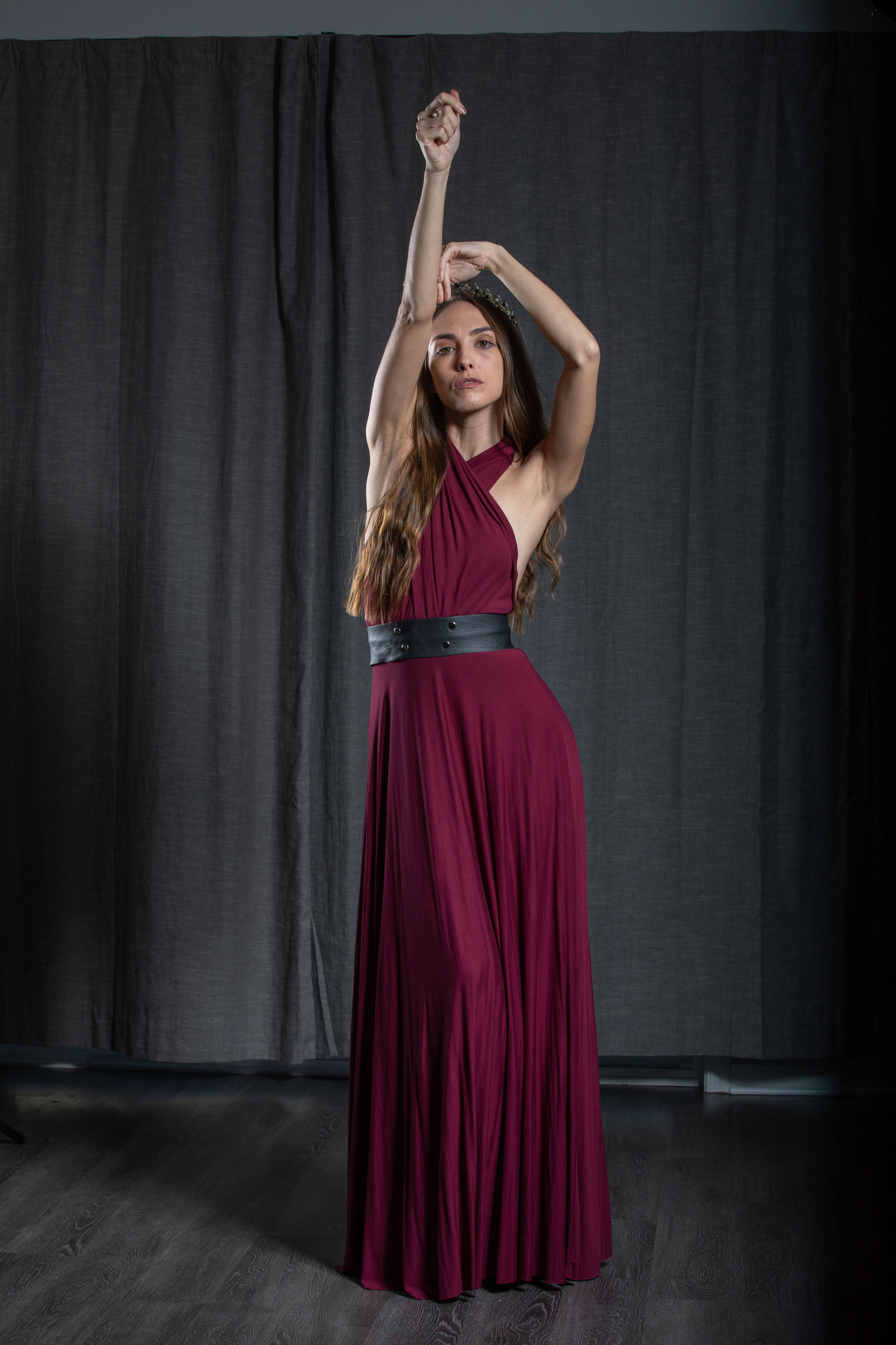 a woman in a long, flowing maroon dress strikes a graceful pose in a studio setting. she has long brown hair and is wearing a black belt that accentuates her waist. the background is a dark gray curtain, and the lighting is soft and diffused, creating a dramatic and elegant atmosphere.
