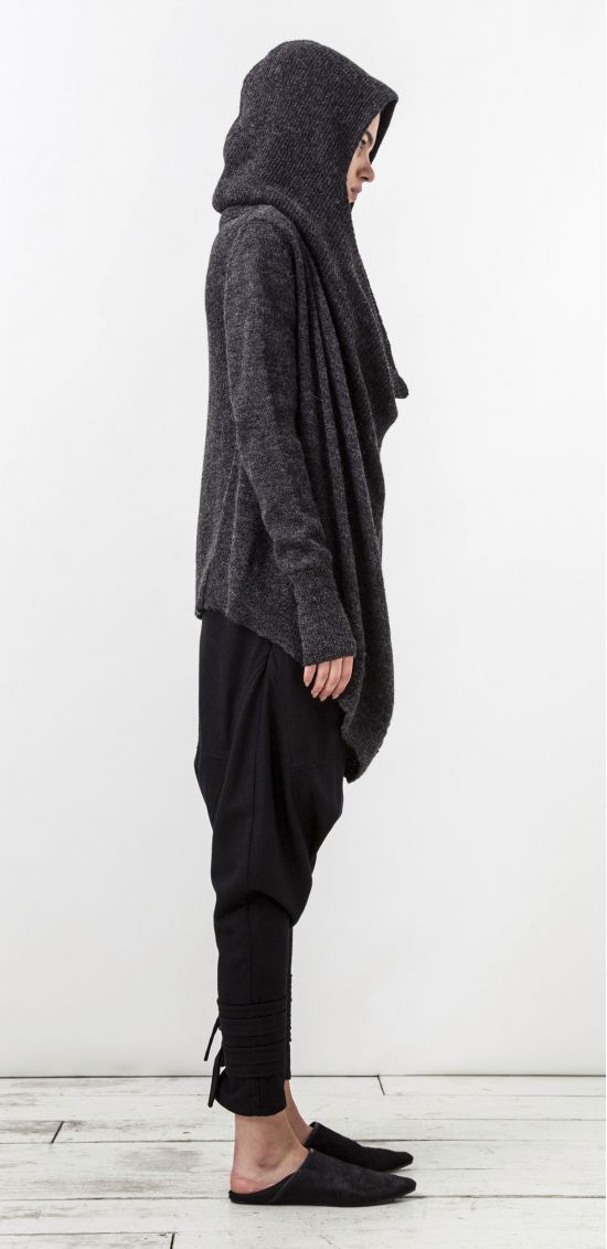 a woman is seen in a side profile, wearing a dark gray, draped cardigan with a hood, paired with black drawstring pants. the pants have a gathered detail at the ankles. she is wearing black mules. the background is a plain, light gray wall.