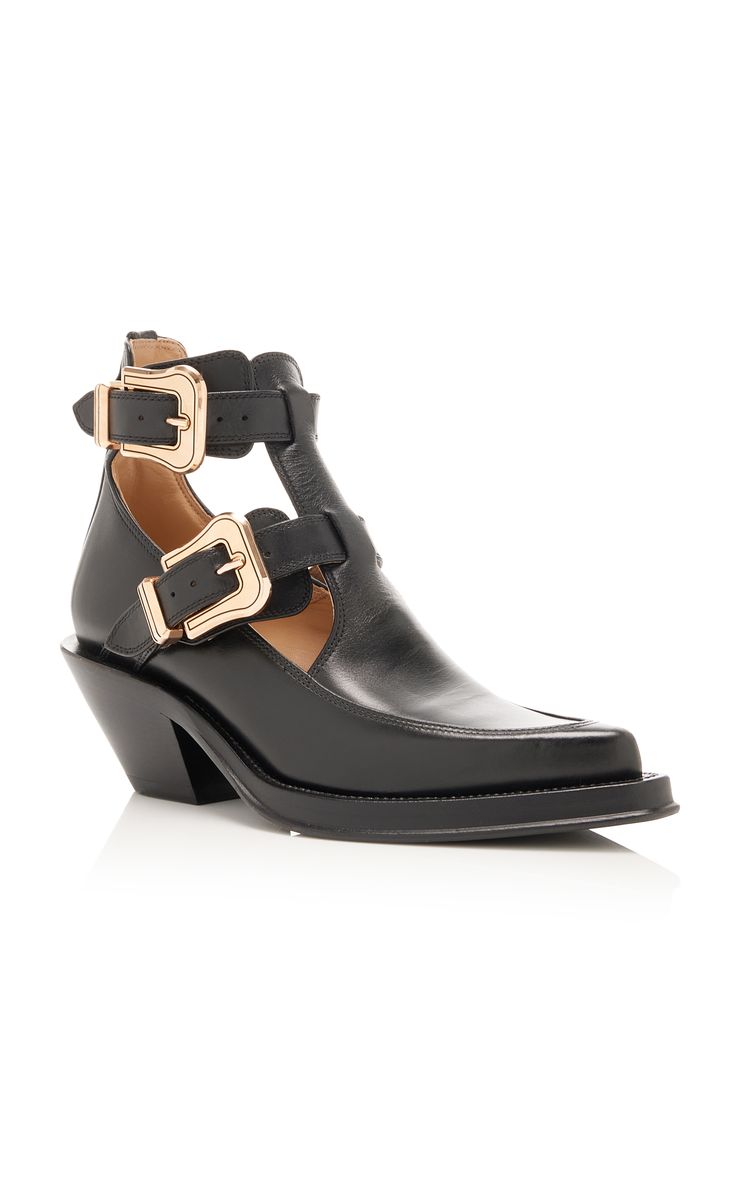 the image shows a pair of women's black leather ankle boots. they are westernstyle boots with a pointed toe and a block heel.  the boots feature a distinctive cutout design on the sides and are adorned with two gold buckles on each ankle strap.  the boots are presented against a clean white background, allowing the details of the shoes to be clearly seen.
