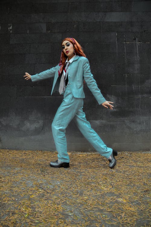 a full shot of a woman in a teal pantsuit walking confidently against a dark grey wall. the background is subtly textured and looks like a wall of large stones. fallen autumn leaves are scattered on the ground. the woman has vibrant red hair tied back and is wearing dark shoes. her expression is one of determined confidence.  the overall aesthetic is stylish and sophisticated.