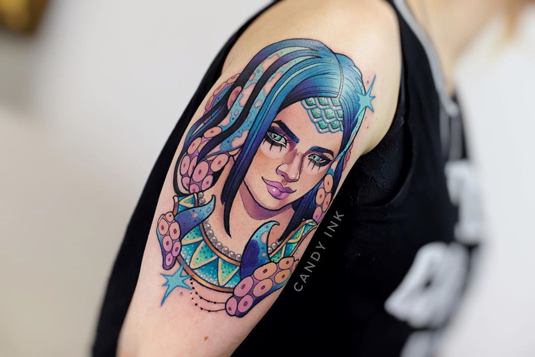 a colorful tattoo of a woman with blue hair and tentacles on the upper arm. the woman is wearing a teal and gold headdress and necklace. the tattoo is done in a cartoon style and is very detailed. the background is black and the tattoo artist's signature, "candy ink", is visible in white in the bottom right corner.