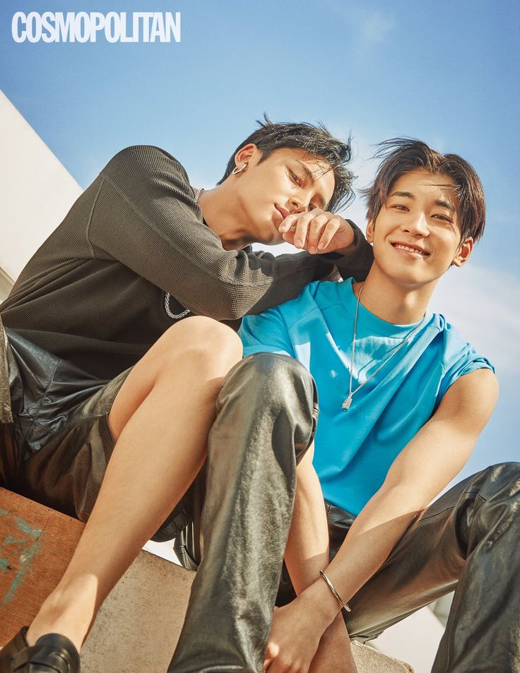 two young men are posing for a magazine photoshoot outdoors. they are sitting on a ledge, smiling and looking at the camera. the man on the left is wearing a black shirt and leather pants, while the man on the right is wearing a blue shirt and leather pants. the sky is blue and there is a lot of sunshine.