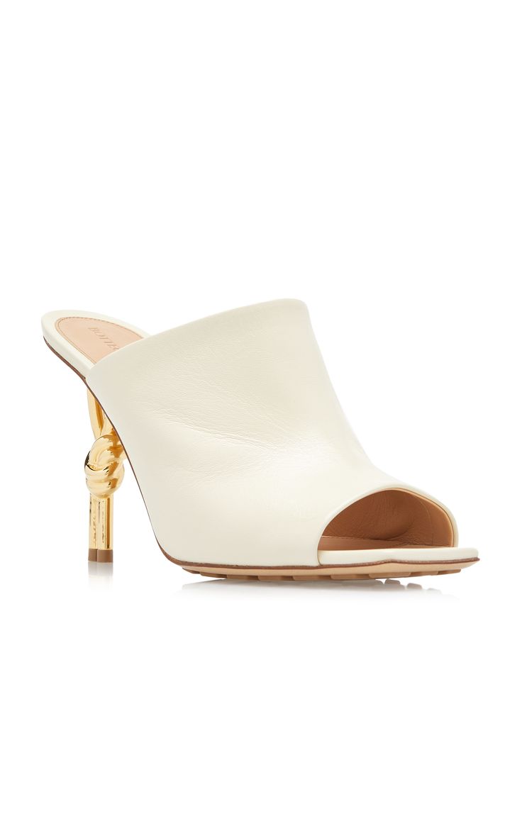 a pair of elegant white leather mules with gold knotshaped heels. the shoes are shown on a white background. the design is minimalist and chic, with a simple silhouette and a unique heel detail. the shoes are perfect for a variety of occasions, from a night out to a wedding.