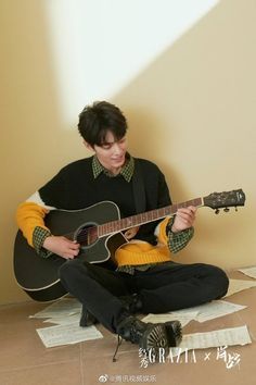 a young asian man sits on a floor scattered with sheet music, playing a black acoustic guitar. he is wearing a black and yellow striped sweater, black jeans, and black boots. the background is a creamcolored wall with a warm light source casting a shadow behind him. the image evokes a sense of peacefulness and creativity.