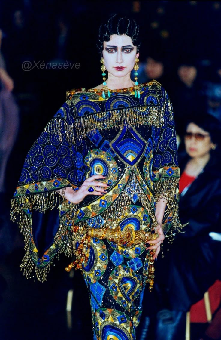 a fashion model poses in an ornate, flowing dress. the woman has dark hair styled in an updo and bold makeup including dramatic dark eyeliner and red lipstick. she is wearing long, dangling earrings and a large ring on her hand. the dress is a vibrant mix of blues, golds, and blacks, heavily embellished with intricate patterns, beading, and long fringe. the patterns include paisley shapes and geometric designs. a decorative gold belt cinches the waist. in the background, a blurred audience can be seen, indicating this is likely a runway show or fashion event. the overall image has a dark and dramatic tone, emphasizing the elaborate detail of the dress and the model's striking appearance.