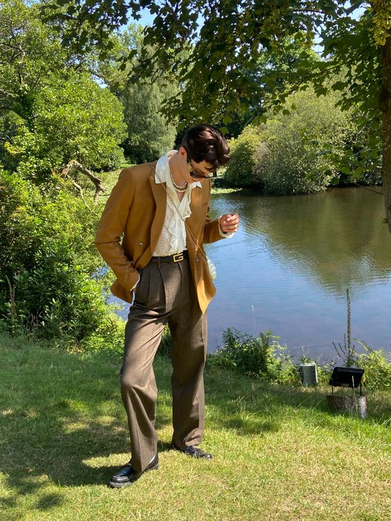 a man in a brown blazer, brown pants, black shoes, and sunglasses is standing in a grassy park by a pond. he is looking down at something off camera. the trees and water are a beautiful backdrop.