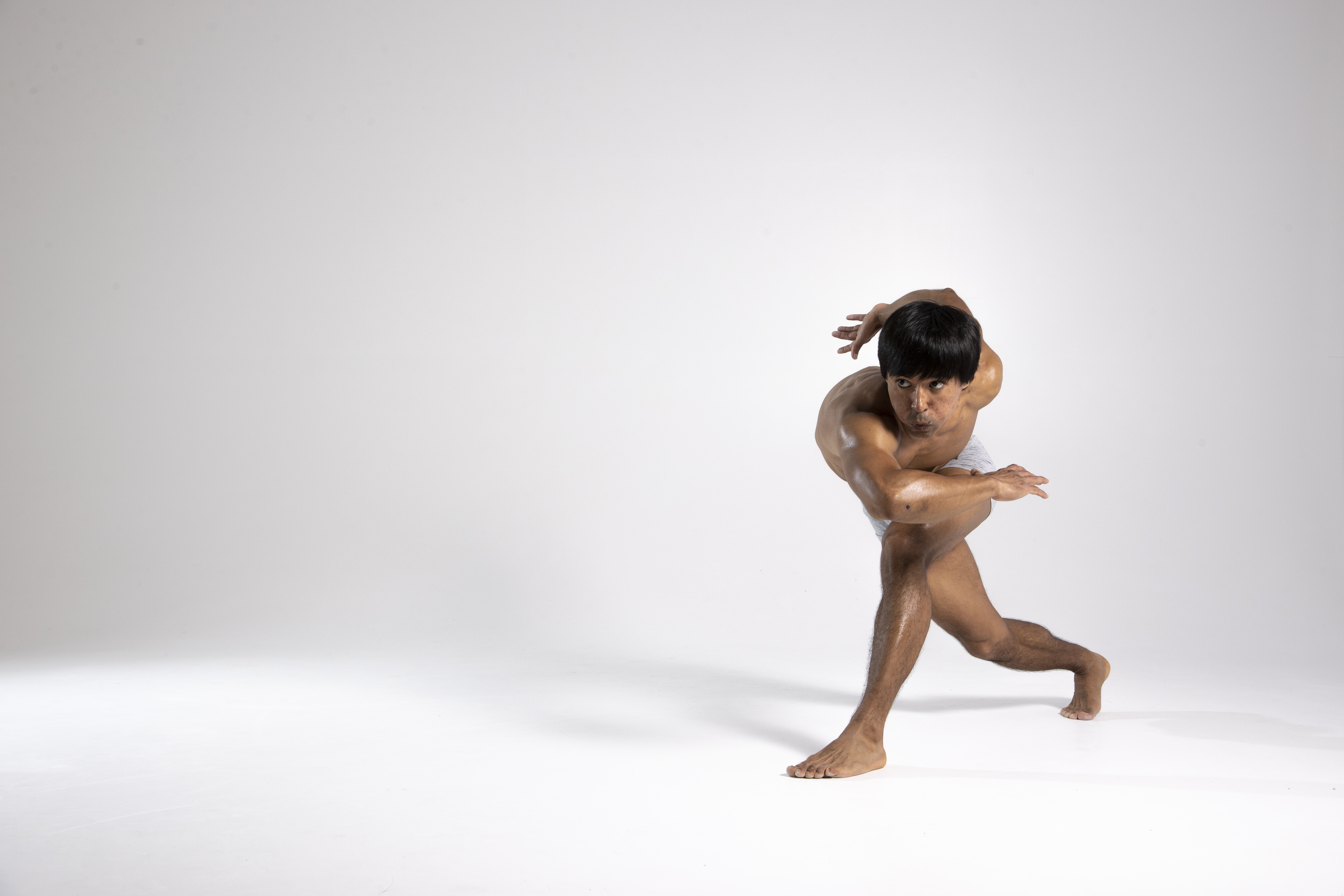 a barechested, muscular man with dark hair is posed in a dynamic, contorted dance position against a white studio background. his body is twisted, with one leg forward and arms crossed in front of him. he appears to be in midmotion, exhibiting athletic prowess, flexibility, and concentration. the image captures a moment of artistic expression and fluid movement.