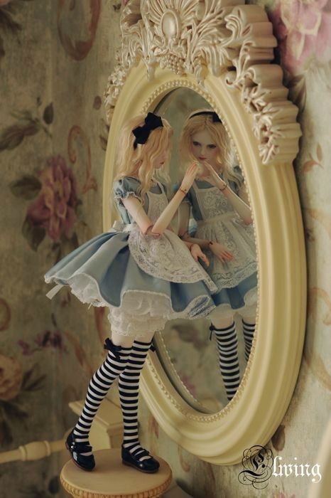 a closeup shot of a doll dressed as alice in wonderland, standing in front of an antique mirror, reflecting a similar doll in a vintage dress with black and white stockings. the doll has blonde hair and wears a blue and white dress with lace accents, and is looking at her reflection in the mirror. the ornate mirror has a creamcolored frame with intricate carvings.  the background has a vintage wallpaper with floral patterns.